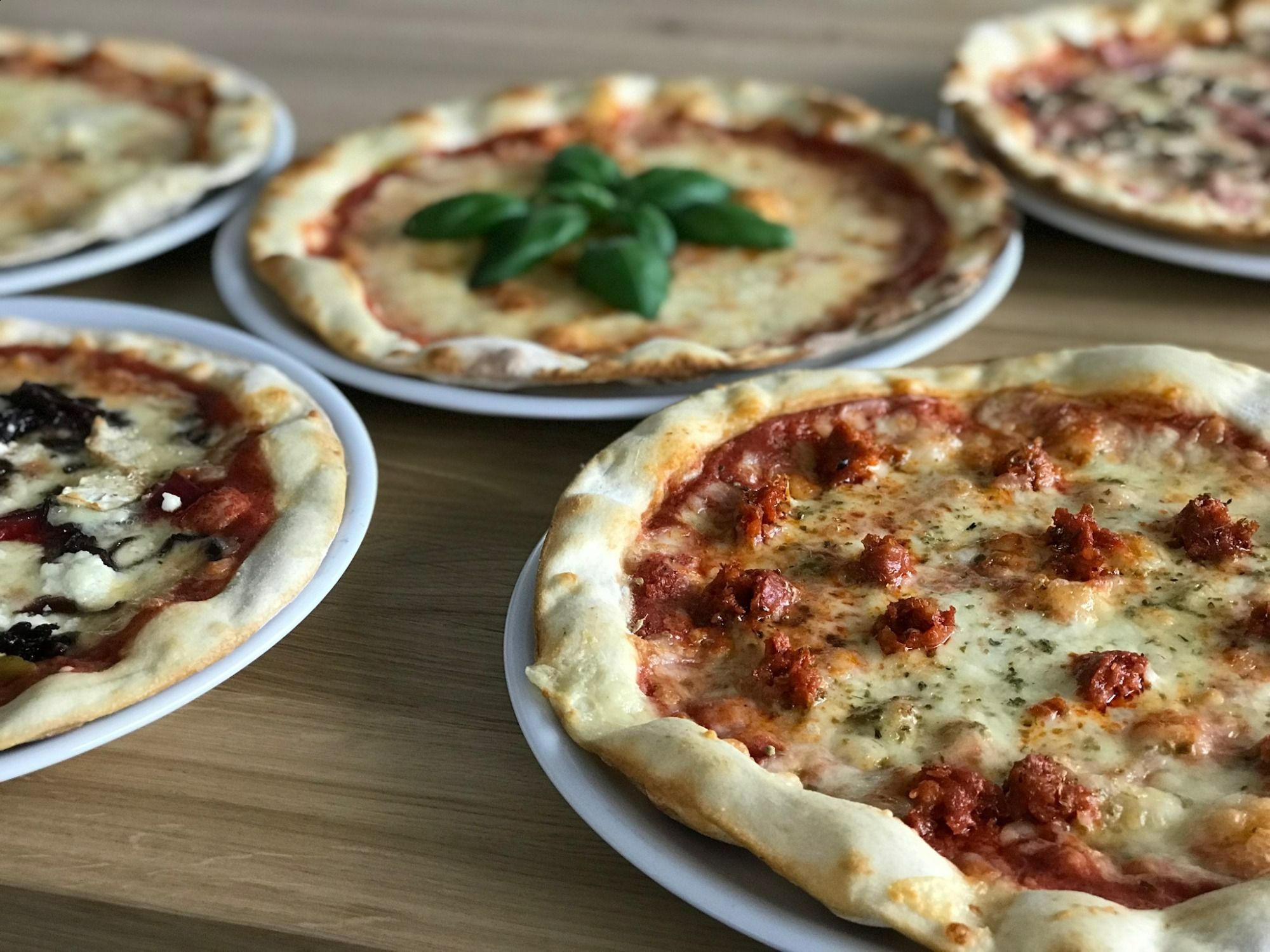 Różne rodzaje pizzy na talerzach, w tym margherita, z kiełbasą i ziołami, na drewnianym stole.