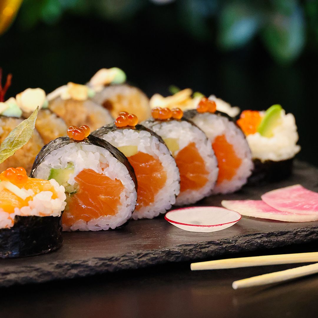 Talerz sushi z rolkami z łososiem, ogórkiem i ikrą, podany na czarnej desce, w tle zielone liście.