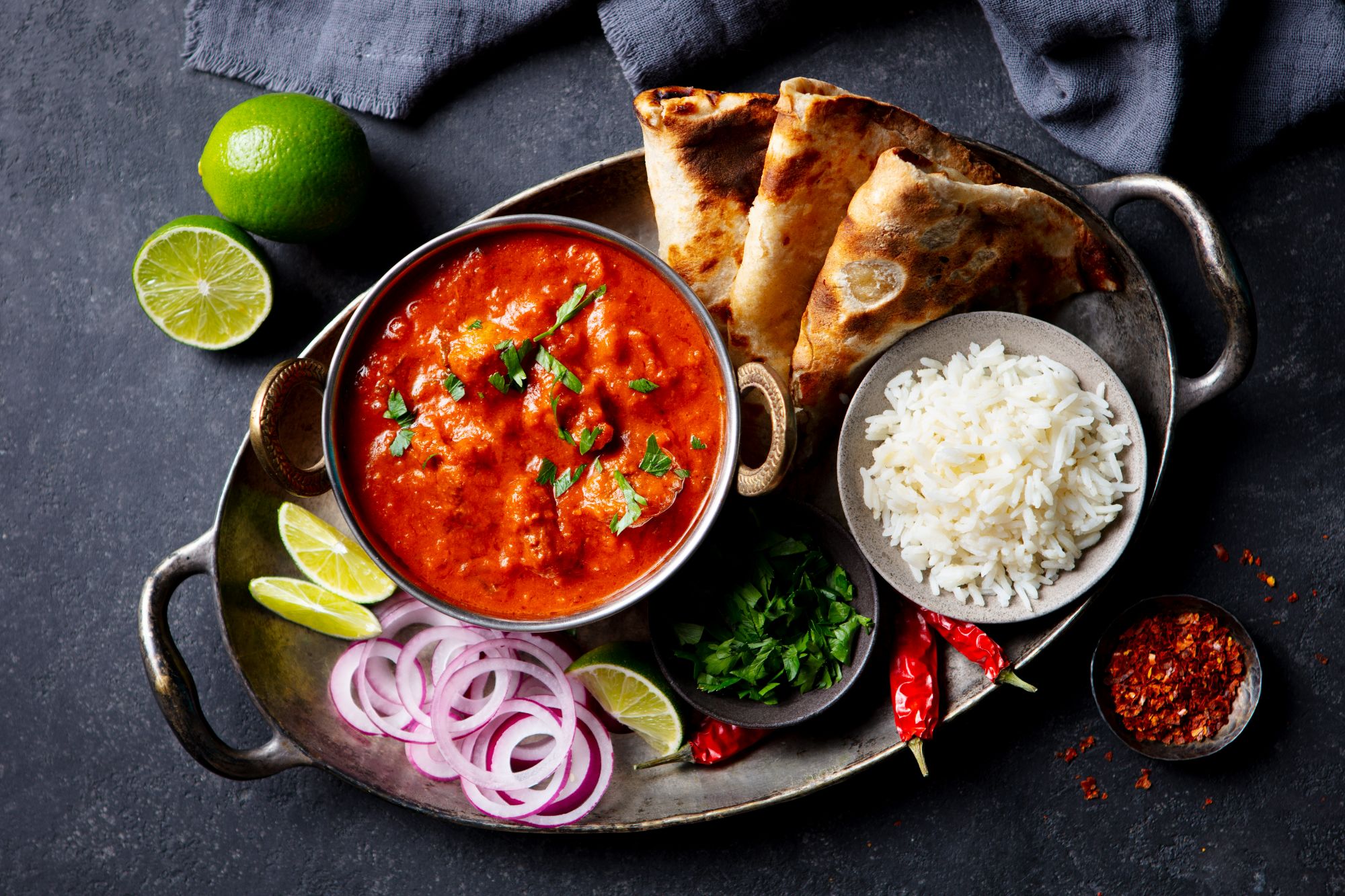 Talerz z curry, ryżem, naan, cebulą, limonką i przyprawami na ciemnym tle.