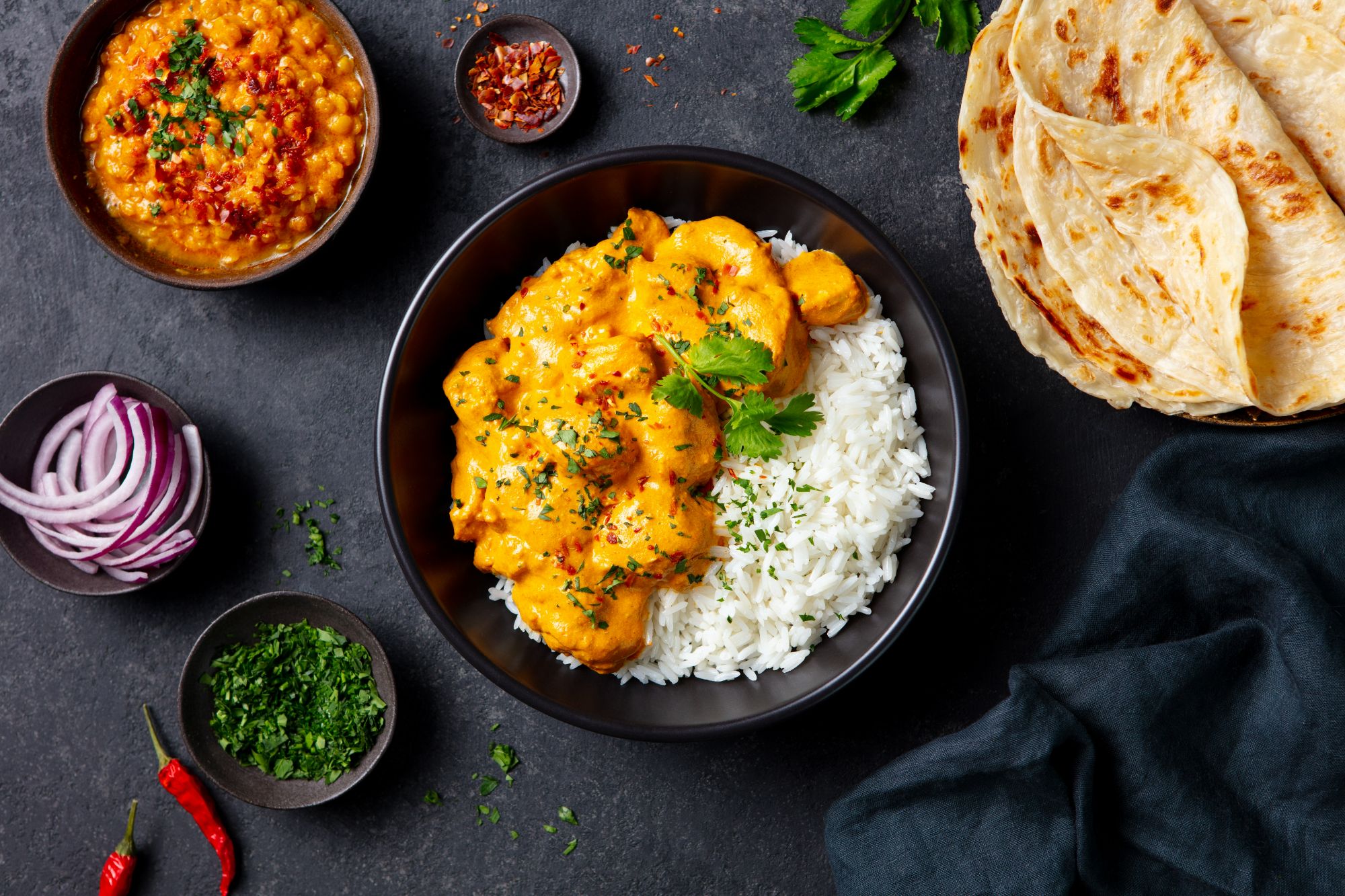 Zestaw dań: curry z kurczaka na ryżu, chlebki naan, cebula, kolendra i przyprawy na ciemnym tle.