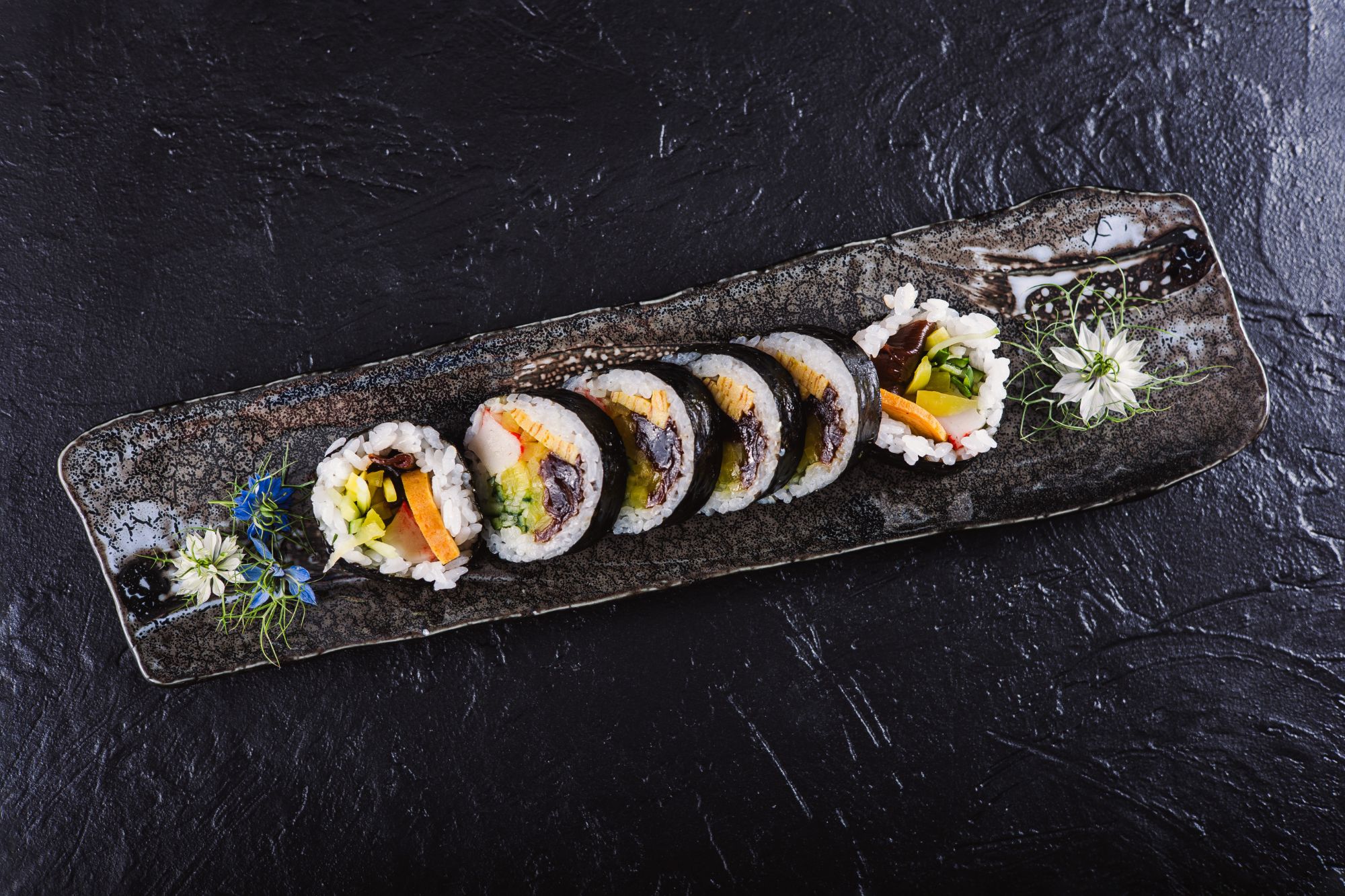 Sushi na talerzu, kawałki z widocznymi warzywami, na czarnym tle.