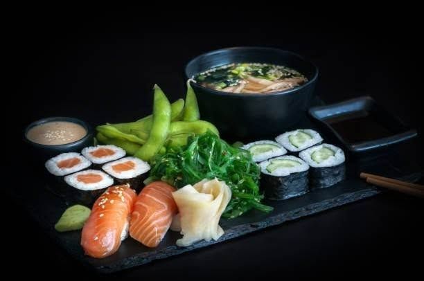Sushi, łosoś, maki z ogórkiem, sałatka wakame, edamame i miska ramen na czarnym tle.