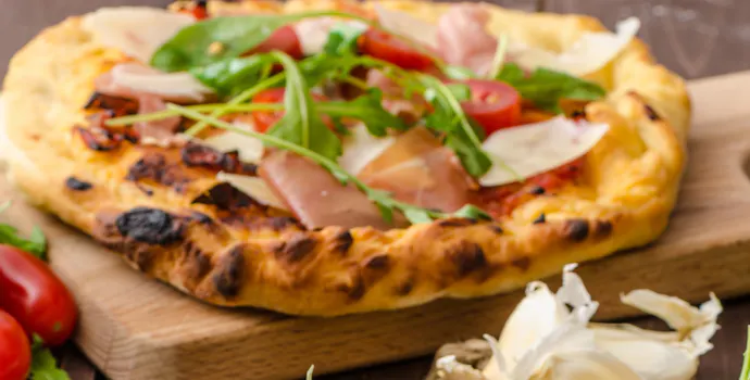 Pizza z rukolą, szynką parmeńską i pomidorkami na drewnianej desce, obok świeże zioła i czosnek.