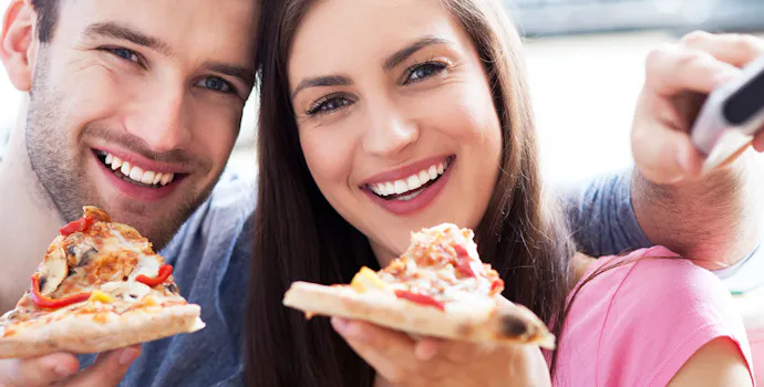 Uśmiechnięta para trzyma pizzę, ciesząc się wspólnym posiłkiem w jasnym wnętrzu.